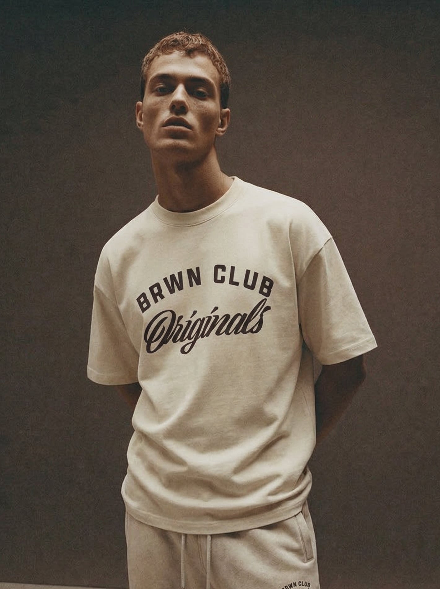 Man wearing a 'Brwn Club Originals' t-shirt and pants against a dark background
